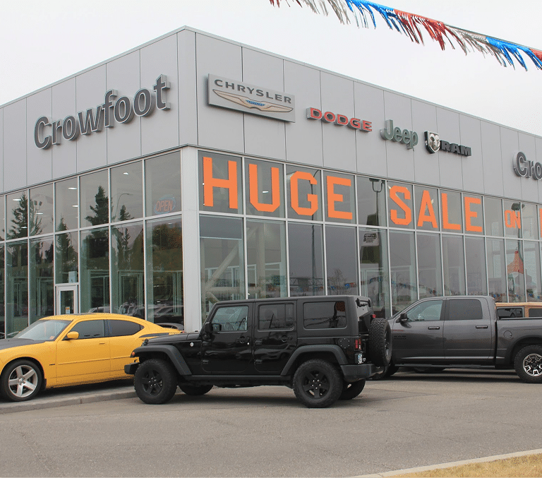 Car dealership with signs for Chrysler, Dodge, Jeep, Ram, and a "Huge Sale" banner. Cars parked in front.