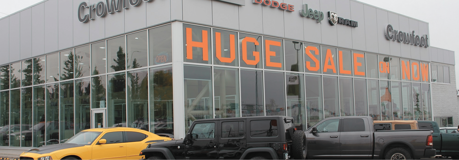Car dealership with large "HUGE SALE ON NOW" sign on windows, featuring cars in the foreground.