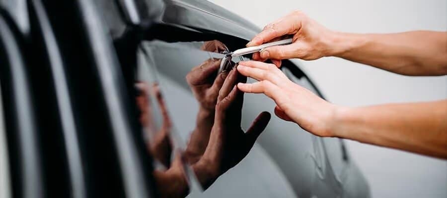 Hands applying window tint film to a car, using a tool for precision installation.
