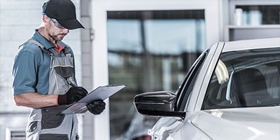 Mechanic in uniform inspects a car, holding a clipboard near a service garage.