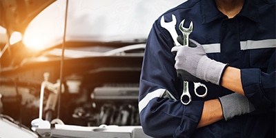 Auto mechanic holding wrenches near an open car hood, sunlight in the background.