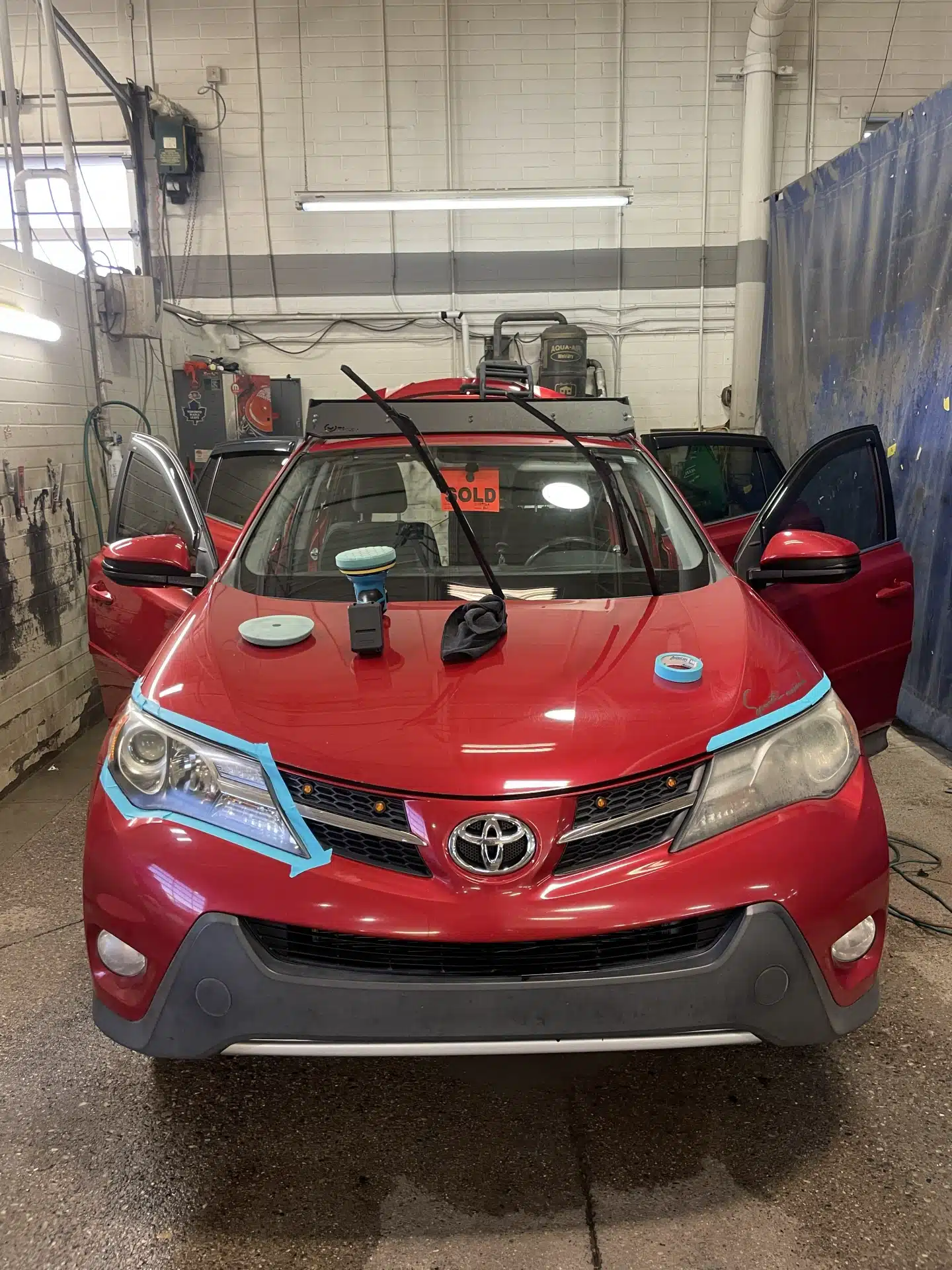 Red Toyota SUV in garage with open doors, detailing tools on hood, and "SOLD" sign on windshield.