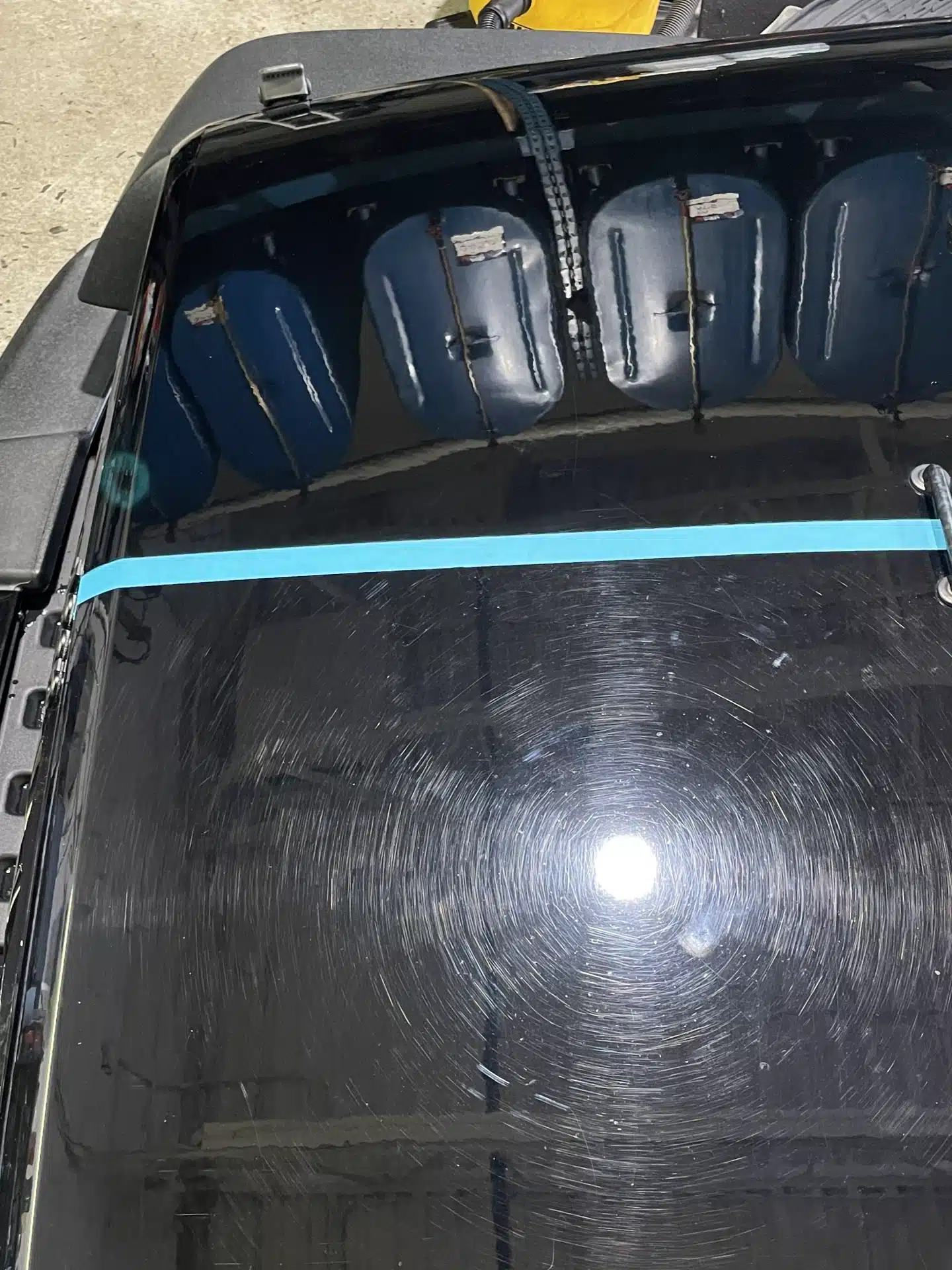 Swirl marks on a car's black paint, with reflections of overhead lights and blue barrels nearby.