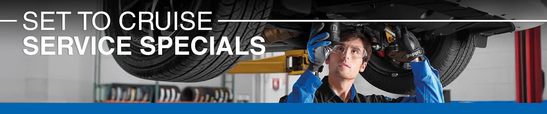 Mechanic in blue uniform examines car undercarriage. Text: "Set to Cruise - Service Specials."